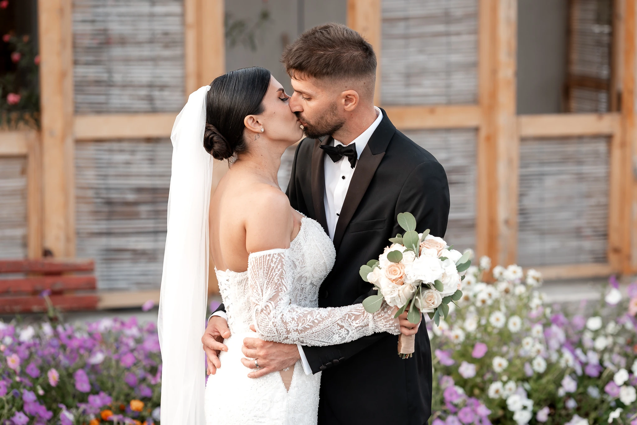 Mireasa Mara și mirele Ionuț se sărută lângă flori și o fațadă din lemn